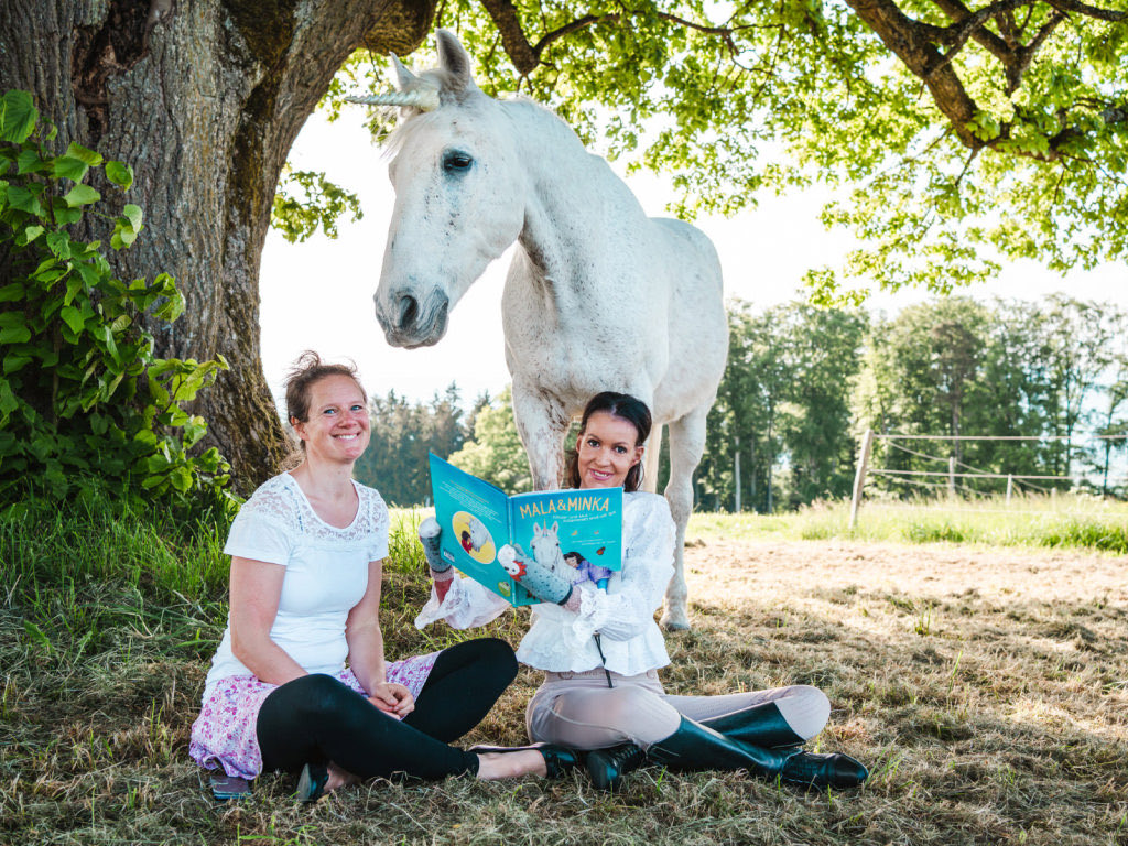 Michelle Zimmermann liest mit zwei Kindern auf einer Parkbank im Kinderbuch „Mala & Minka“; hinter der Bank steht ein weisses Pony mit Einhorn-Horn.